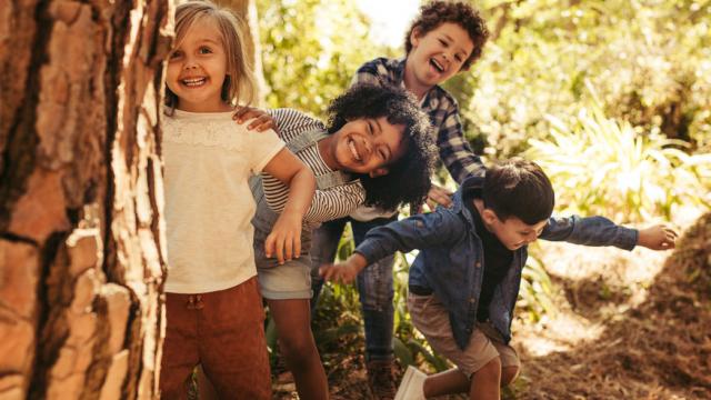 Cute smiling kids peeking out from behind the tree in the park. Group of children enjoying playing hide and seek in a forest.
